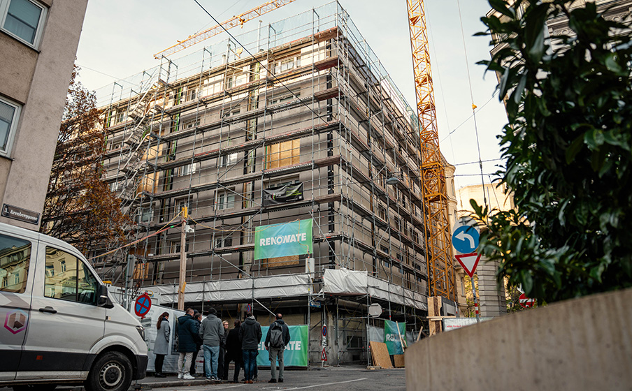 Multi-story building wrapped in scaffolding and 'RENOWATE' banners, construction crane overhead, van and group of people on the street.