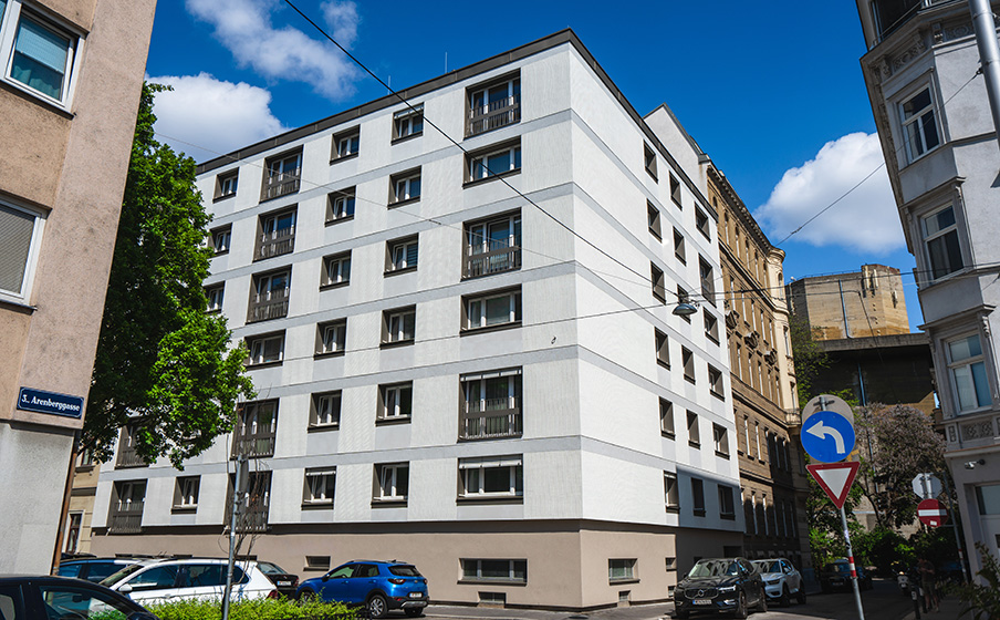 White multi-story building on a sunny day, surrounded by parked cars and trees, with a street sign in the foreground.