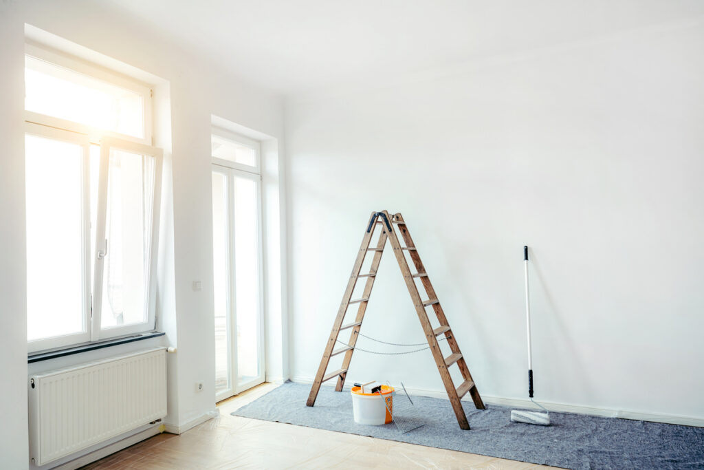 Ein Wohnraum mit großen Fenstern und einer Leiter, dabenen ein Eimer mit Farbe und eine Malerrolle